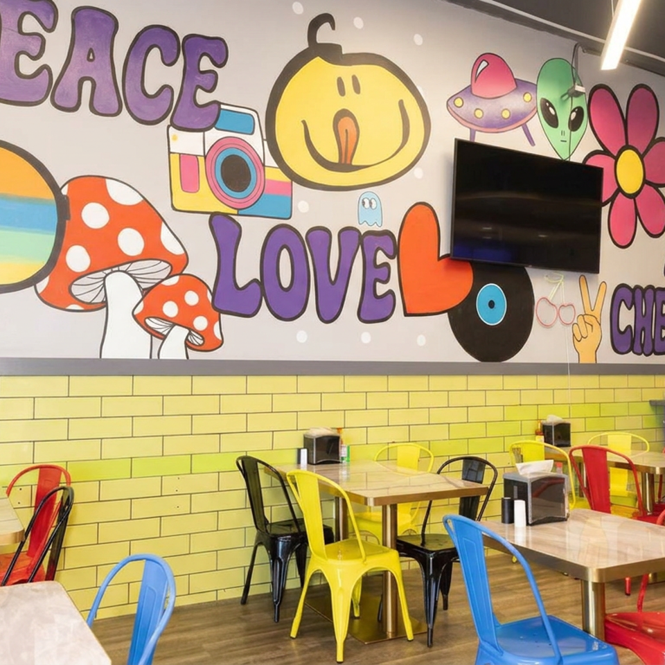 Colorful restaurant dining area with multicolored metal chairs, wooden tables, yellow brick walls, and a playful alien-themed mural.